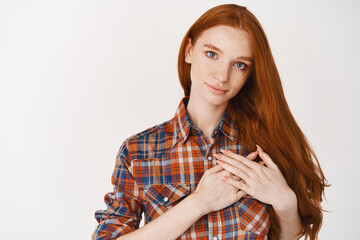 Naklejka premium Young caring and heartfelt woman holding hands on heart, thanking for something, standing over white background