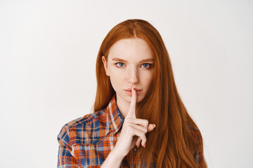 Serious young lady with red natural hair, shushing at camera, telling a secret with confident face, standing over white background