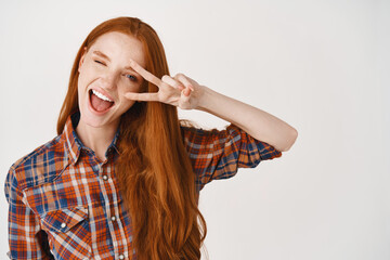 Beautiful redhead female showing peace sign and winking, smiling happy at camera, standing over white background