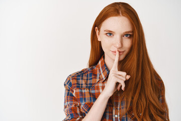 Beautiful smiling woman with long ginger hair and blue eyes, sharing a secret, hushing with finger on lips, standing over white background