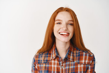 Young attractive redhead girl showing tongue, smiling with white teeth, looking carefree at camera, standing over white background