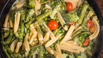 Cooking primavera pasta in a pan - vegan american and italian primavera pasta dish with broccoli, beans, asparagus, peas and tomatoes in a pan on a wooden table, top view