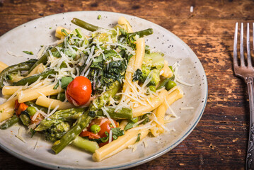 Vegetarian american and italian primavera pasta dish with broccoli, beans, asparagus, peas and tomatoes in a plate on a wooden table, top view