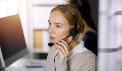 Businesswoman talking by headset while sitting in sunny office. Call center and diverse people...