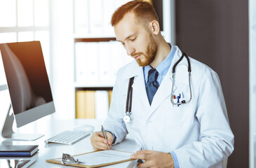 Friendly red-bearded doctor sitting and writing at clipboard in sunny clinic. Medicine concept