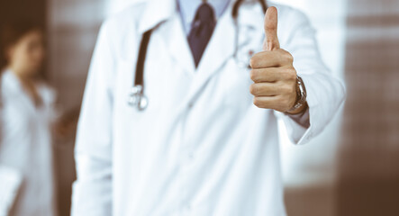 Unknown man-doctor standing straight and showing Ok sign with thumb up in modern clinic. Female colleague is in a hurry at the background of physician. Medicine concept