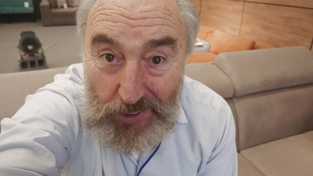 Chest-up shot of 60-something Caucasian man with cropped grey hair sitting on couch at home, holding invisible smartphone in outstretched hand, waving and chatting enthusiastically on video call
