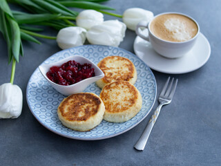 Syrniki, curd or cottage cheese pancakes with cherry jam and coffee on a grey background With white tulips, top view. Healthy diet breakfast