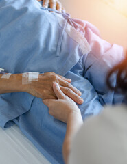 Caregiver and elderly senior patient (aged old adult person) holding hands in hospital bed or...