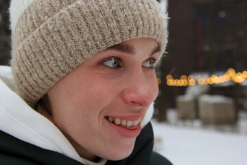 Close-up of the face of a smiling blue-eyed girl in a winter hat against the background of a winter park and lights