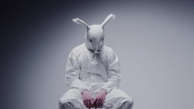 Weird, strange person in an animal mask shaking on a chair in a white lab room. White rabbit, mad man. Hallucination on drugs, crazy vision in a dream. Mental disorder, creepy craziness and paranoia.