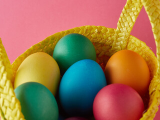 Fototapeta na wymiar Yellow straw basket with multi colored eggs on pink background.