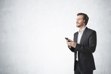Young European man in elegant suit listening to music with headphones and smartphone, concrete