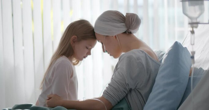 Side view of young woman with cancer sitting on hospital bed with little daughter