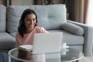 Smiling indian female freelancer wearing headset working in comfort living room look at pc screen talking by video call. Young arabian woman study from home on distance take part in conference webinar