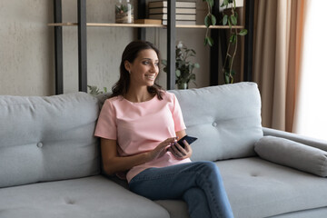 Smiling indian lady spend good weekend at home check social network read news using modern cell look aside enjoy leisure time. Young arabian woman sit on sofa feel glad engaged in easy shopping online