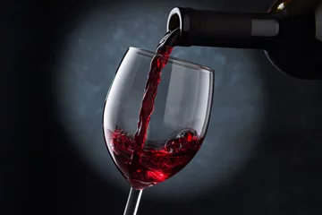 Fotobehang Rode wijn wordt in een glas gegoten uit een fles op een wazige blauwe achtergrond, een stroom rode wijn uit de fles wervelt in het glas, close-up. Vrije ruimte voor tekst. © Александр Кузьмин