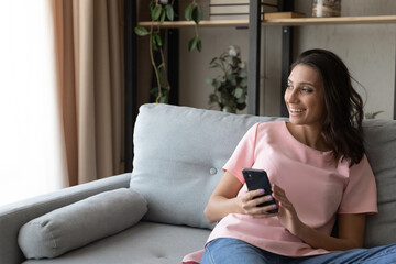 Young arab woman sit on comfy couch look at window with smile distracted from cell screen get...