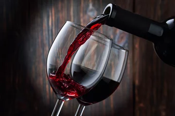 Fotobehang Rode wijn wordt in een glas gegoten uit een fles op een wazige houten achtergrond, een stroom rode wijn uit de fles wervelt in het glas, close-up. Vrije ruimte voor tekst. © Александр Кузьмин