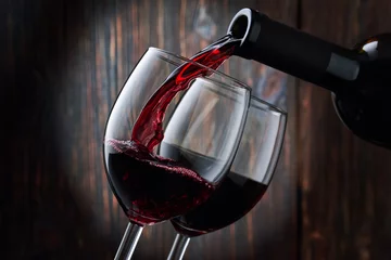 Fotobehang Red wine is poured into a glass from a bottle on a blurred wooden background, a stream of red wine from the bottle swirls in the glass, close-up. Free space for text. © Александр Кузьмин