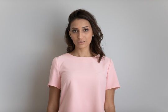 Studio portrait of serious beautiful young indian arabian female with long hair wearing pink blouse. Head shot of millennial pretty mixed race female looking at camera posing on light grey background