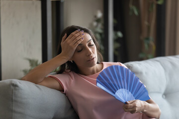Too hot. Exhausted indian female suffer from heat in modern apartment use hand fan feel lack of air conditioning. Unhappy young mixed race female complaining on high temperature at home hard to breath