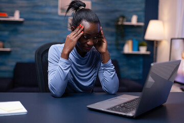 Dark skinned woman touching foarhead because of headache while working from home. Tired focused employee using modern technology network wireless doing overtime.