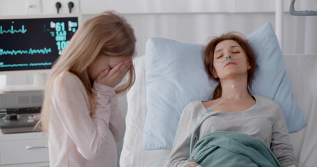 Little girl crying standing near sick mother sleeping in hospital bed