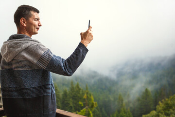 Young man takes photos with his phone of a beautiful view from the hotel's balcony overlooking the mountains. Mountain view from the window of fir trees, fog and hills in the home. Selfie. Copy space