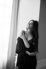 Attractive young girl with long hair in jeans and a black shirt stands at a large window 