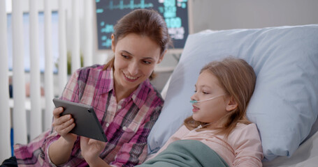 Mother and daughter in hospital bed watching cartoons on digital tablet