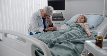Fototapeta premium Worried aged doctor holding hand of sick child sleeping in hospital bed