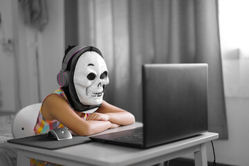 Asian child or kid girl funny e-learning on computer notebook with wearing headphone and white ghost mask for video call communication and study online or learn from home by play laptop back to school