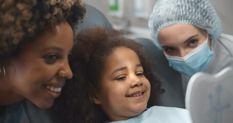 Cute kid visiting dentist with mom sitting in dentist chair and looking in mirror at beautiful smile - Powered by Adobe