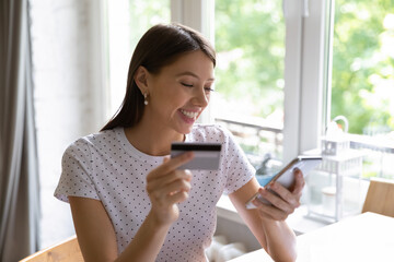 Smiling young Caucasian woman look at cellphone screen shopping online paying with credit card. Happy millennial female buyer or client buy on internet, make payment on smartphone with secure service.