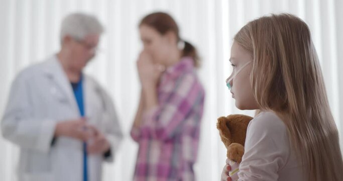 Sad sick kid with teddy bear sitting in hospital bed with doctor talking to upset mom on background