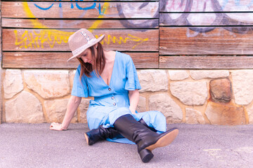 Beautiful middle-aged young woman sitting on the pavement floor with stone and wooden background wearing hat, leather boots and blue dress. spring summer fashion concept