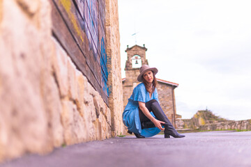 Obraz na płótnie Canvas Beautiful middle-aged young woman sitting on the pavement floor with stone and wooden background wearing hat, leather boots and blue dress. spring summer fashion concept