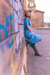 Beautiful middle-aged young woman leaning on a stone and wooden wall with hat, leather boots and blue dress. spring summer fashion concept