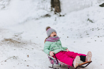 A beautiful girl of preschool age, a child, dressed in warm clothes, in a frosty winter goes down the hill on a wooden, metal sled on fresh white snow. Emotional photography.