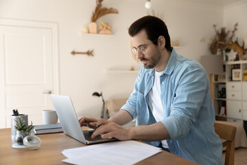 Serious millennial Caucasian businessman sit at desk at home office work online on laptop with documents. Pensive young man look at computer screen busy engaged in distant job, consulting client.