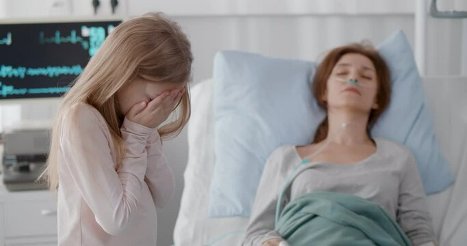 Little girl crying standing near sick mother sleeping in hospital bed