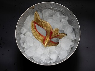 Traditional golden carnival Venetian mask with red ornaments in a bucket with crushed ice cubes. Brazilian Mardi Gras Festival. The concept of a hot parade, masquerade.