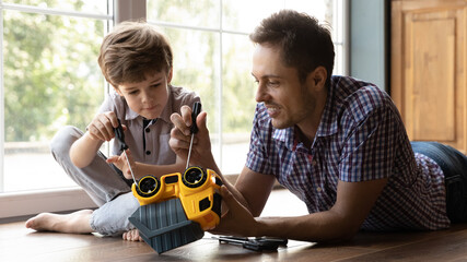 Close up caring father and little son fixing toy car, lying on warm wooden floor at home, smiling...