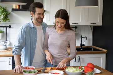 Happy millennial Caucasian man and woman spouses have fun cooking healthy diet salad for dinner. Smiling young couple renters enjoy morning at home preparing delicious vegetarian food together.