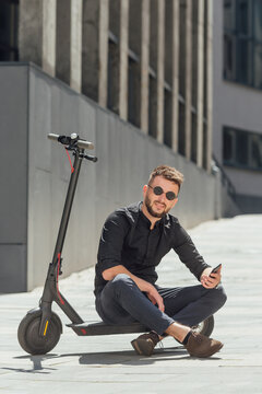 Young Man Sitting On  Electric Scooter With Phone In Urban Background