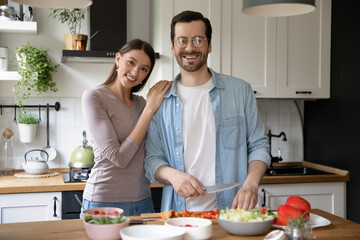 Obraz na płótnie Canvas Portrait of happy young family pose in modern cozy home kitchen preparing healthy vegetarian salad. Smiling millennial couple renters cooking delicious diet food in the morning in own modern house.