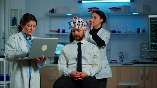 Medic in neuroscience working in neurological research laboratory developing brain experiment holding laptop explaning to man brainwave scanning headset side effects of nervous system treamtment.