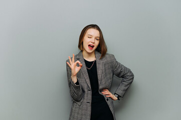 Emotional happy office woman in jacket smiling looking at camera, winking and showing OK gesture,...