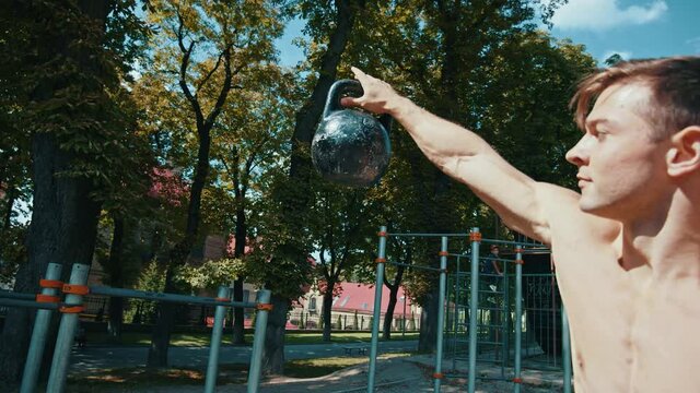 Side view of strong athletic man training with weight at street. Caucasian guy throwing a sports kettlebell from hand to hand while exercising at sunny summer day. Sport and recreation concept